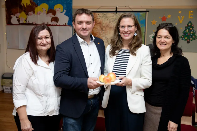 Vier Personen stehen eng beieinander in einem Raum. Ein Mann und eine Frau halten einen Teller mit Äpfeln. Die Frau trägt eine Brille und einen weißen Blazer. Dahinter befindet sich eine Wand mit einer Karte und einem Banner.