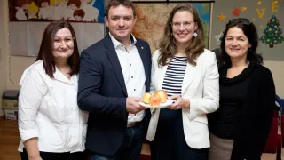 Vier Personen stehen eng beieinander in einem Raum. Ein Mann und eine Frau halten einen Teller mit Äpfeln. Die Frau trägt eine Brille und einen weißen Blazer. Dahinter befindet sich eine Wand mit einer Karte und einem Banner.