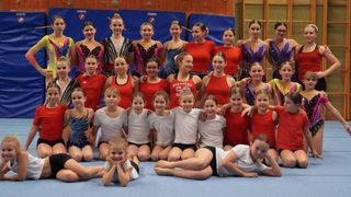 Eine Gruppe von Turnerinnen in roten und weißen Trikots posiert für ein Foto. Sie sind in verschiedenen Posen, einige knien, andere stehen und manche sitzen. Hinter ihnen ist eine blaue Wand mit der Aufschrift 'NIKE PRO'. An den Seiten befinden sich auch Holzpaneele.