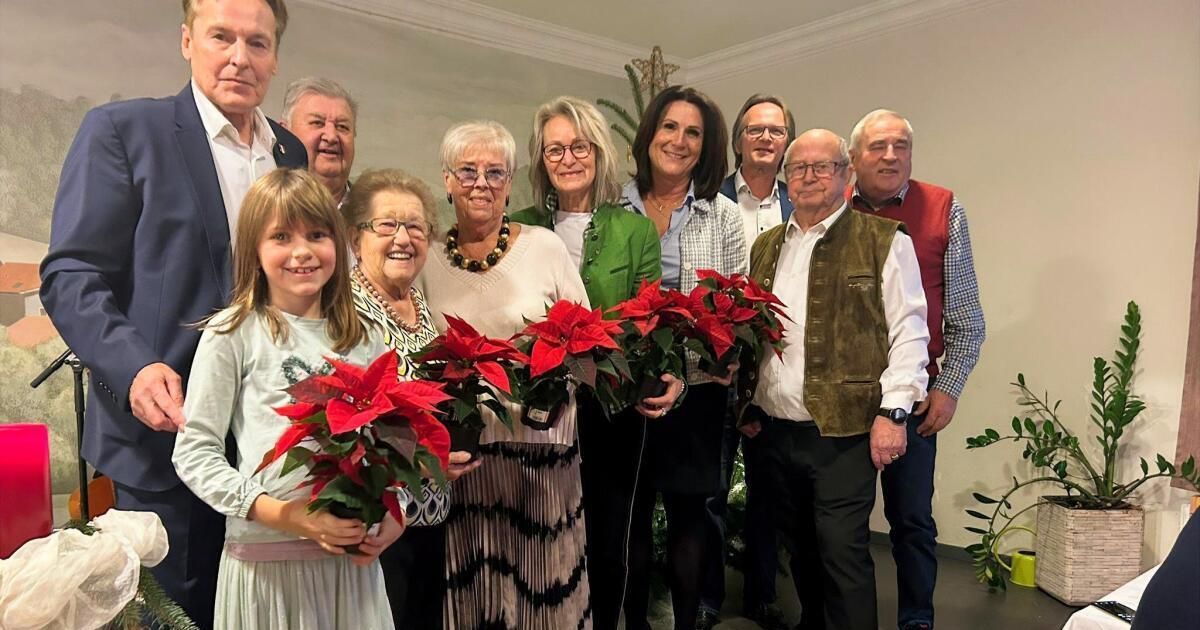 Eine Gruppe von Menschen, einschließlich Kinder, stehen lächelnd für ein Foto, während sie rote Weihnachtssterne halten. Sie sind in einem Raum mit einem Weihnachtsbaum und weißen Wänden.