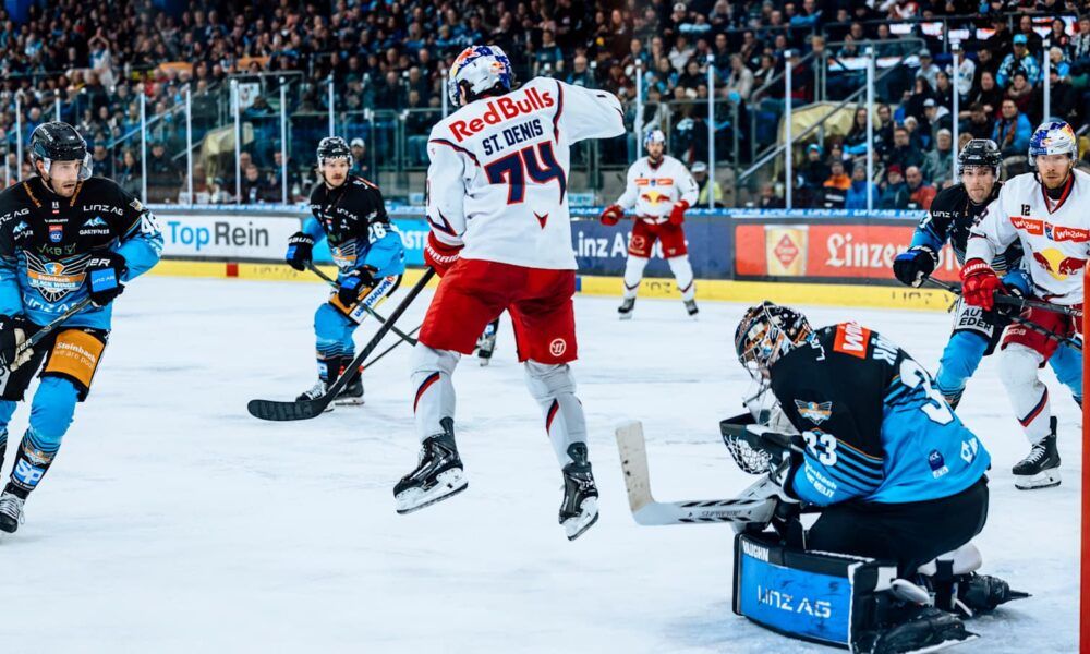 Eishockeyspieler in Aktion auf einer Eisbahn. Ein Spieler in einem Red Bull-Trikot versucht zu treffen. Der Torwart ist gekauert und verteidigt das Tor.