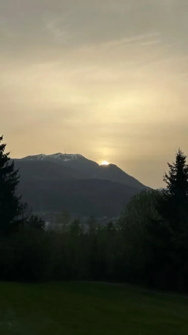 Die Sonne geht hinter einem Gebirgszug unter und taucht die Landschaft in ein warmes Licht. Im Vordergrund sind Bäume und Büsche zu sehen.