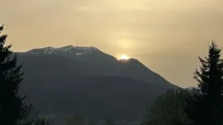 Die Sonne geht hinter einem Gebirgszug unter und taucht die Landschaft in ein warmes Licht. Im Vordergrund sind Bäume und Büsche zu sehen.
