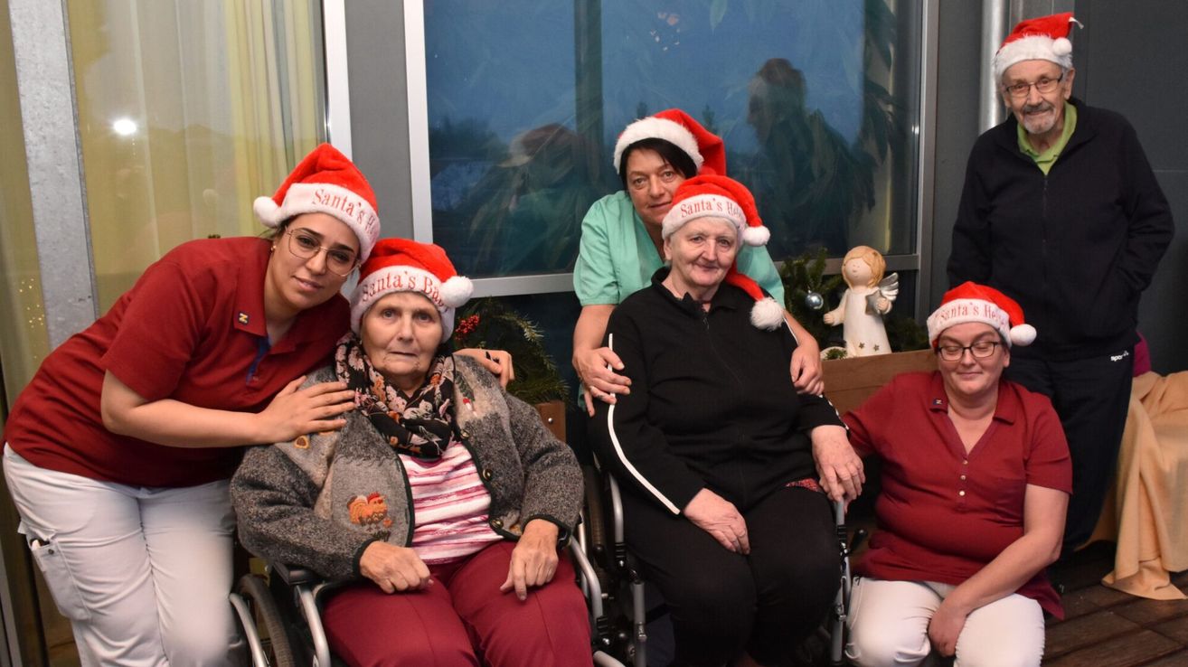 Eine Gruppe älterer Frauen und eine Krankenschwester posieren für ein Foto. Sie tragen Weihnachtsmützen, sitzen in Rollstühlen und haben einen Weihnachtsbaum im Hintergrund.
