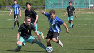 Vier Fußballspieler auf einem Feld. Ein Spieler in Blau läuft mit dem Ball, während ein anderer in Schwarz versucht, ihn zu stehlen. Zwei weitere beobachten von hinten.