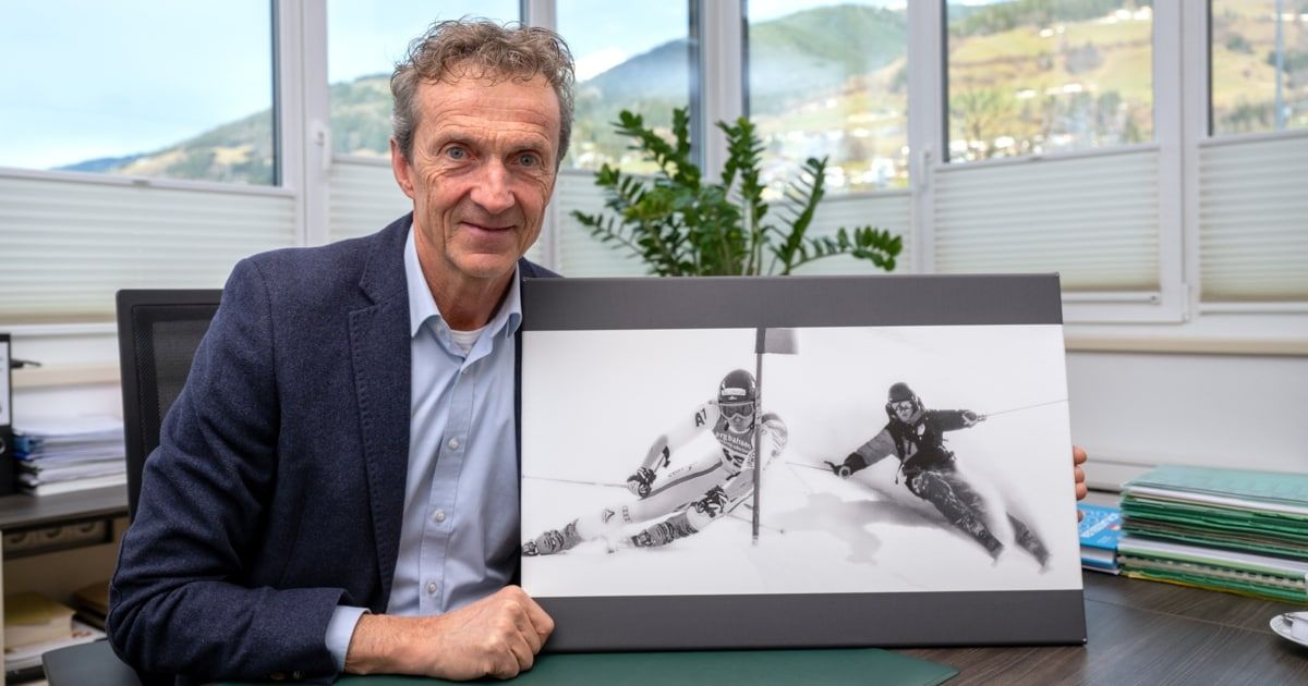 Ein Mann lächelt, während er ein gerahmtes Foto von zwei Skifahrern hält, einer im Vordergrund und einer im Hintergrund, mit Bergen und einem Fenster hinter ihm.