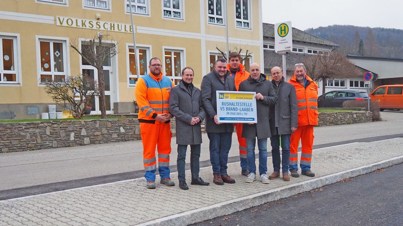 Fünf Männer in Warnwesten stehen auf einem Bürgersteig und halten ein blaues Schild, wahrscheinlich bei einer Gemeinschaftsveranstaltung. Hinter ihnen ist ein Gebäude mit großen Fenstern zu sehen.