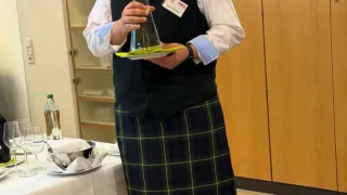 Eine Person in einem schwarzen Weste und einer karierten Schürze, die eine große Glasvase in einem Raum mit Tisch und Schränken hält.