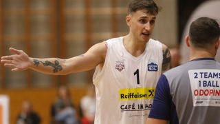 Ein Basketballspieler mit weißem Trikot, Nummer 1 und Kreuz-Tätowierung spricht mit einem anderen Spieler. Das Trikot hat Raiffeisen-, Rocks- und Austria B2L-Logos.