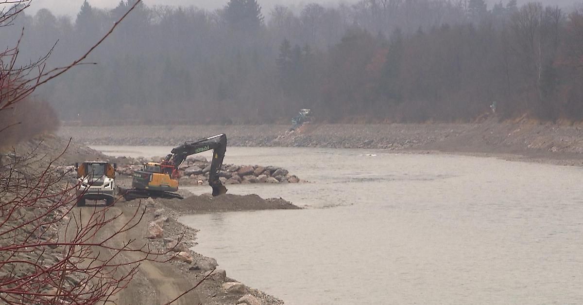 Eine Baustelle in der Nähe eines Flusses, mit einem Bagger, der in Kies gräbt. Nebel bedeckt die Bäume im Hintergrund.