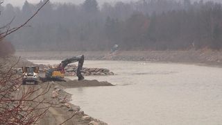 Eine Baustelle in der Nähe eines Flusses, mit einem Bagger, der in Kies gräbt. Nebel bedeckt die Bäume im Hintergrund.