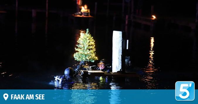 In der Nacht schwimmt ein kleines Boot mit einem leuchtenden Weihnachtsbaum im Wasser. Zwei Menschen tauchen in der Nähe. Ein Steg ist im Hintergrund.