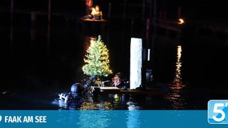 In der Nacht schwimmt ein kleines Boot mit einem leuchtenden Weihnachtsbaum im Wasser. Zwei Menschen tauchen in der Nähe. Ein Steg ist im Hintergrund.