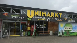 Die Fassade eines Unimarkt Regional-Geschäfts zeigt ein Schild mit gelben Buchstaben, das den Slogan 'Genuss verbindet' trägt. Der Eingang ist mit Mülleimern und einem Coca-Cola-Automaten geschmückt.