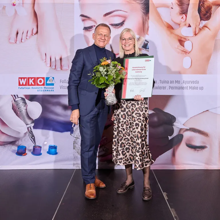 Ein Mann und eine Frau stehen auf der Bühne und halten ein Zertifikat und Blumen. Sie lächeln und die Frau trägt ein Leoparden-Kleid. Hinter ihnen ist eine Kulisse mit Bildern von Schönheitsbehandlungen.