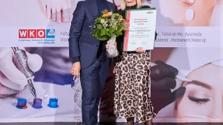 Ein Mann und eine Frau stehen auf der Bühne und halten ein Zertifikat und Blumen. Sie lächeln und die Frau trägt ein Leoparden-Kleid. Hinter ihnen ist eine Kulisse mit Bildern von Schönheitsbehandlungen.