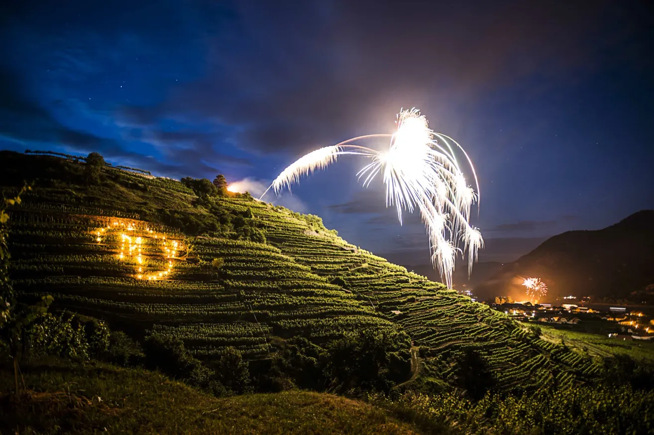 Ein nächtlicher Blick auf terrassierte Weinberge mit Feuerwerken, die den Himmel erhellen. Die Feuerwerke explodieren in einer Farbenpracht und schaffen ein wunderschönes Spektakel. Die Landschaft wird von dem sanften Leuchten der Lichter beleuchtet.