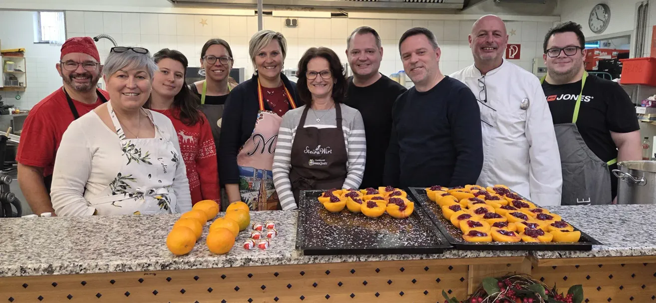 Eine Gruppe von Menschen lächelt in einer Küche. Sie tragen Schürzen und posieren für ein Foto. Vor ihnen steht ein Tisch mit zwei Tabletts mit Essen und Orangen.