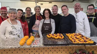 Eine Gruppe von Menschen lächelt in einer Küche. Sie tragen Schürzen und posieren für ein Foto. Vor ihnen steht ein Tisch mit zwei Tabletts mit Essen und Orangen.