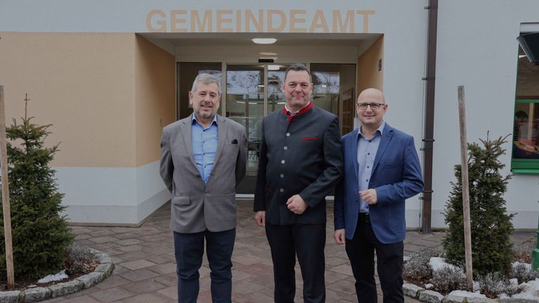 Drei Männer stehen vor einem Gebäude mit dem Schild Gemeindeamt über ihnen. Die Männer sind in Anzügen gekleidet. Der Mann auf der linken Seite trägt eine Brille und hat einen Bart. Der Mann in der Mitte trägt einen schwarzen Anzug. Der Mann auf der rechten Seite ist kahlköpfig.