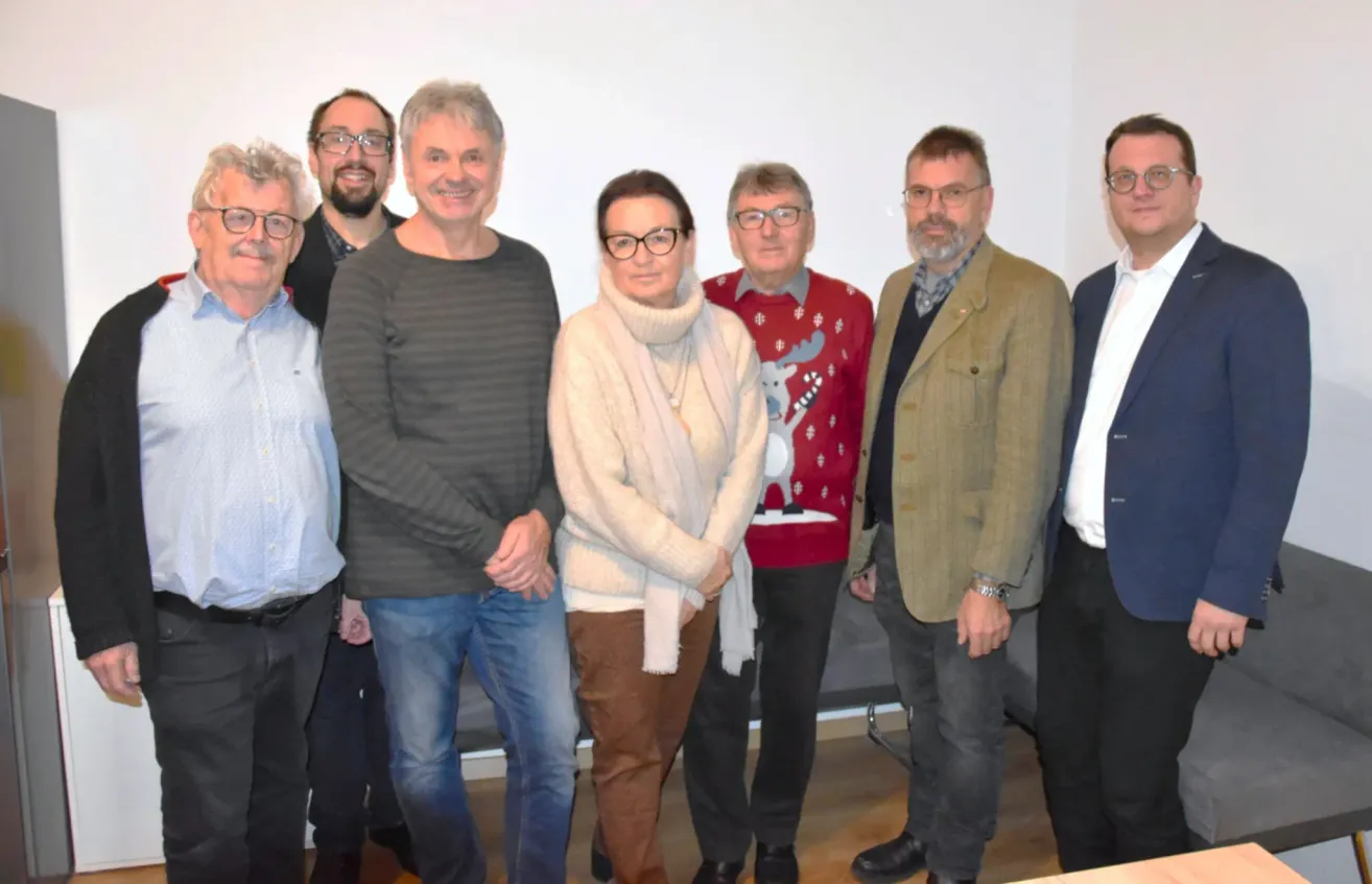 Eine Gruppe von Personen steht in einem Raum zusammen, wahrscheinlich für ein Foto. Sie tragen alle eine Brille und einige tragen eine Uhr. Sie stehen vor einer weißen Wand.