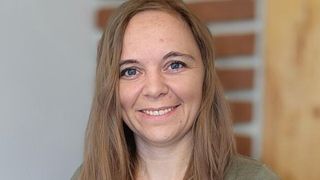 Eine Frau mit langen blonden Haaren lächelt und posiert für ein Foto. Sie hat blaue Augen und einen Muttermal auf ihrer Stirn. Sie trägt ein grünes Oberteil.