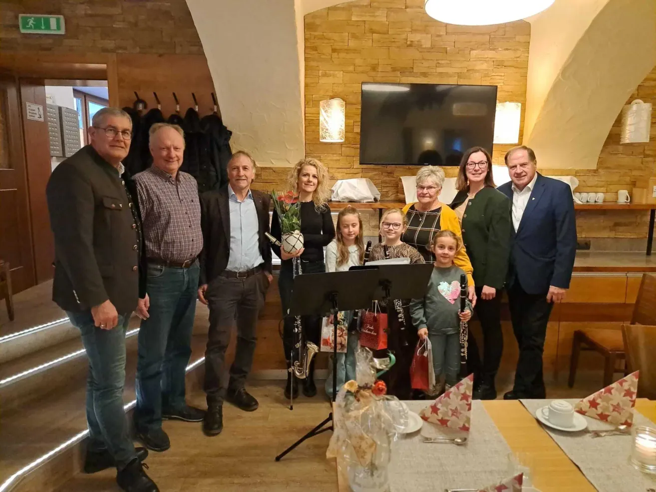 Eine Gruppe von Menschen steht in einem Raum mit Musikinstrumenten, darunter ein Saxophon und eine Trompete. Einige von ihnen lächeln und zwei Kinder halten Geschenke. Es gibt einen Tisch mit einer Blume, einer Tasse und einer Untertasse.