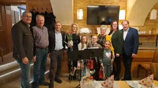 Eine Gruppe von Menschen steht in einem Raum mit Musikinstrumenten, darunter ein Saxophon und eine Trompete. Einige von ihnen lächeln und zwei Kinder halten Geschenke. Es gibt einen Tisch mit einer Blume, einer Tasse und einer Untertasse.