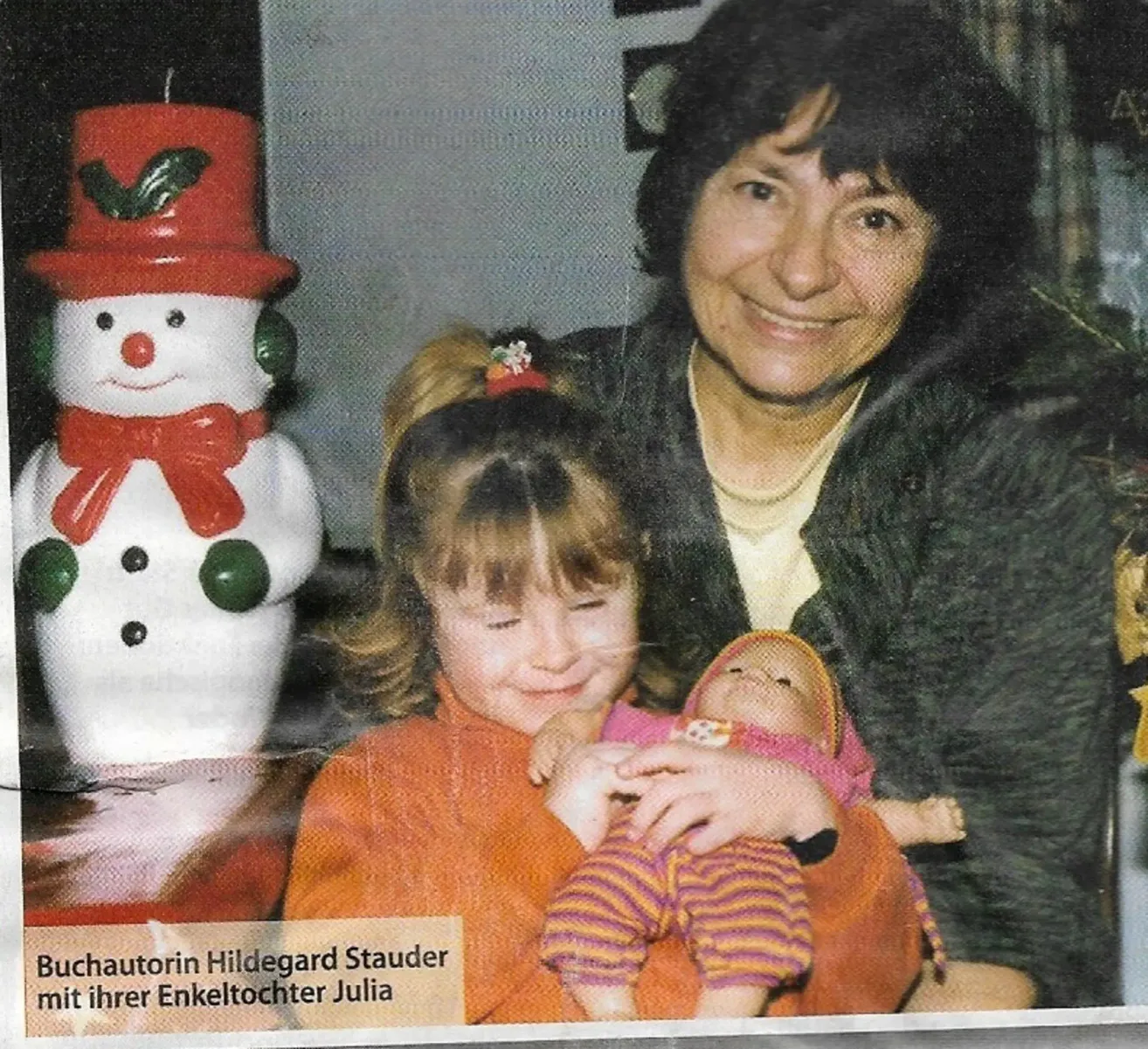 Eine Frau und ein junges Mädchen lächeln und halten eine Babypuppe. Ein Schneemann steht in der Nähe, und ein Bildunterschrift lautet 'Kinderpflegerin Hildegard Stauder mit ihrer Enkeltochter Julia.'