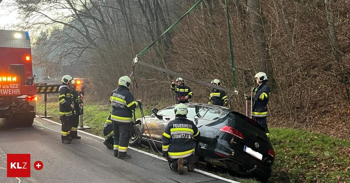 Feuerwehrleute arbeiten daran, ein beschädigtes Auto von der Straße zu extrahieren. Mehrere tragen Helme und Schutzausrüstung. Das Auto ist von der Straße in einen Grasbereich gerutscht.