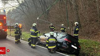 Feuerwehrleute arbeiten daran, ein beschädigtes Auto von der Straße zu extrahieren. Mehrere tragen Helme und Schutzausrüstung. Das Auto ist von der Straße in einen Grasbereich gerutscht.