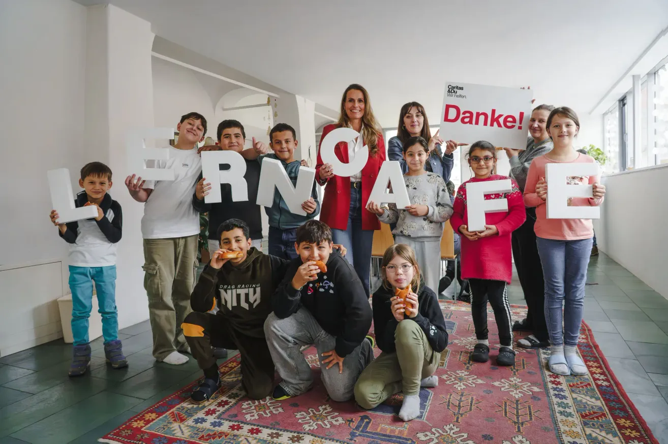 Eine Gruppe von Kindern und Erwachsenen posiert für ein Foto und hält Buchstaben, die 'ESSEN' bilden. Sie scheinen alle etwas zu essen. Hinter ihnen befindet sich ein Schild mit dem Wort 'Danke!' darauf.