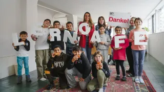 Eine Gruppe von Kindern und Erwachsenen posiert für ein Foto und hält Buchstaben, die 'ESSEN' bilden. Sie scheinen alle etwas zu essen. Hinter ihnen befindet sich ein Schild mit dem Wort 'Danke!' darauf.