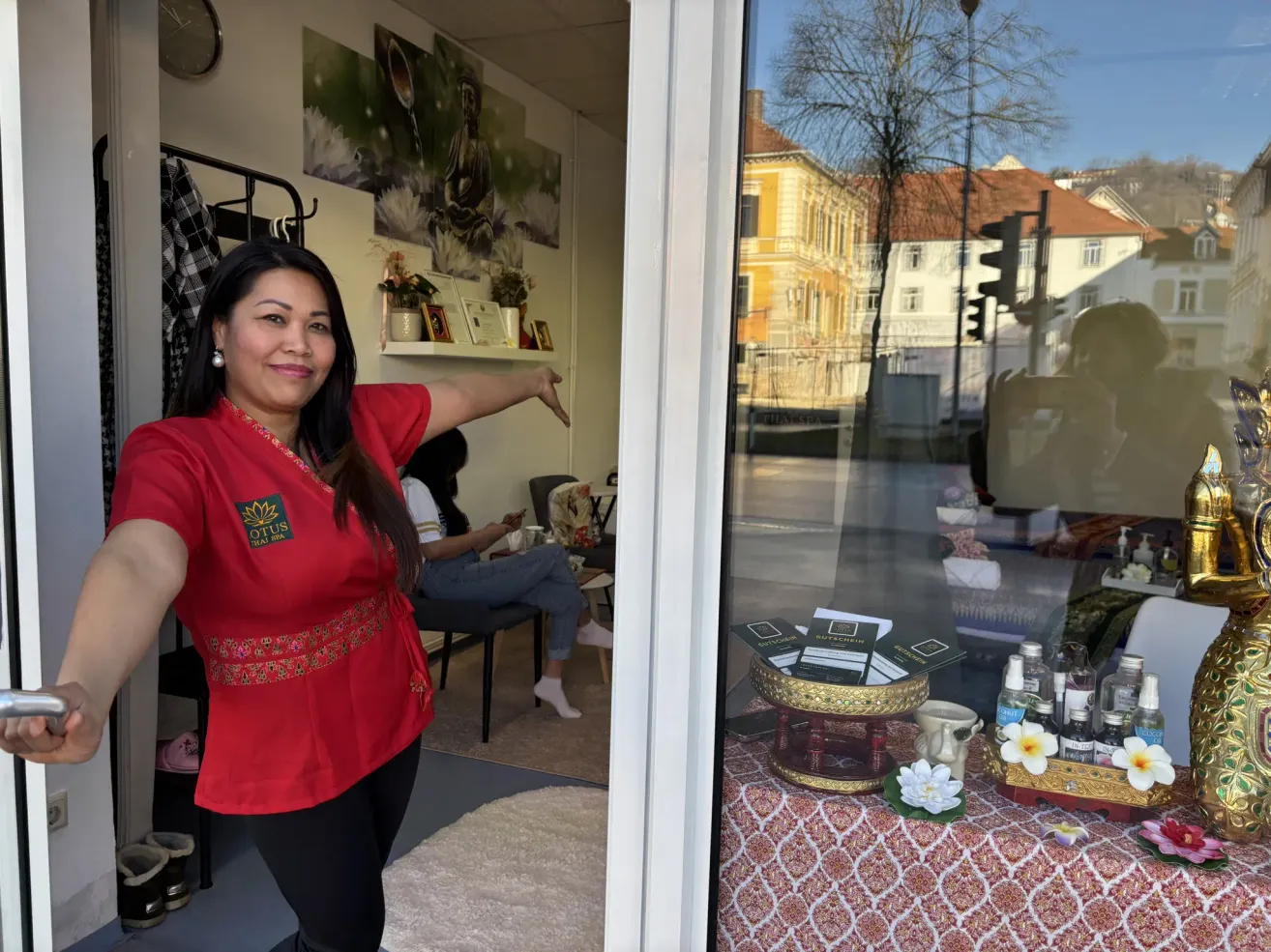 Eine Frau in einem roten Shirt steht neben einer Glastür und zeigt. Im Inneren ist ein Tisch mit Produkten und Blumen zu sehen, mit Gebäuden, die draußen reflektiert sind.