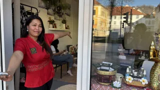 Eine Frau in einem roten Shirt steht neben einer Glastür und zeigt. Im Inneren ist ein Tisch mit Produkten und Blumen zu sehen, mit Gebäuden, die draußen reflektiert sind.