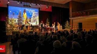 Ein Weihnachtskonzert findet in einer Halle statt. Eine Gruppe von Menschen tritt auf der Bühne mit Weihnachtsdekorationen und einer großen Leinwand hinter ihnen auf. Das Publikum sitzt vor der Bühne.