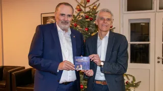 Zwei Männer stehen vor einem Weihnachtsbaum, einer hält eine Karte und der andere lächelt. Der Mann auf der linken Seite hält eine Karte mit einem Bild von zwei Personen.