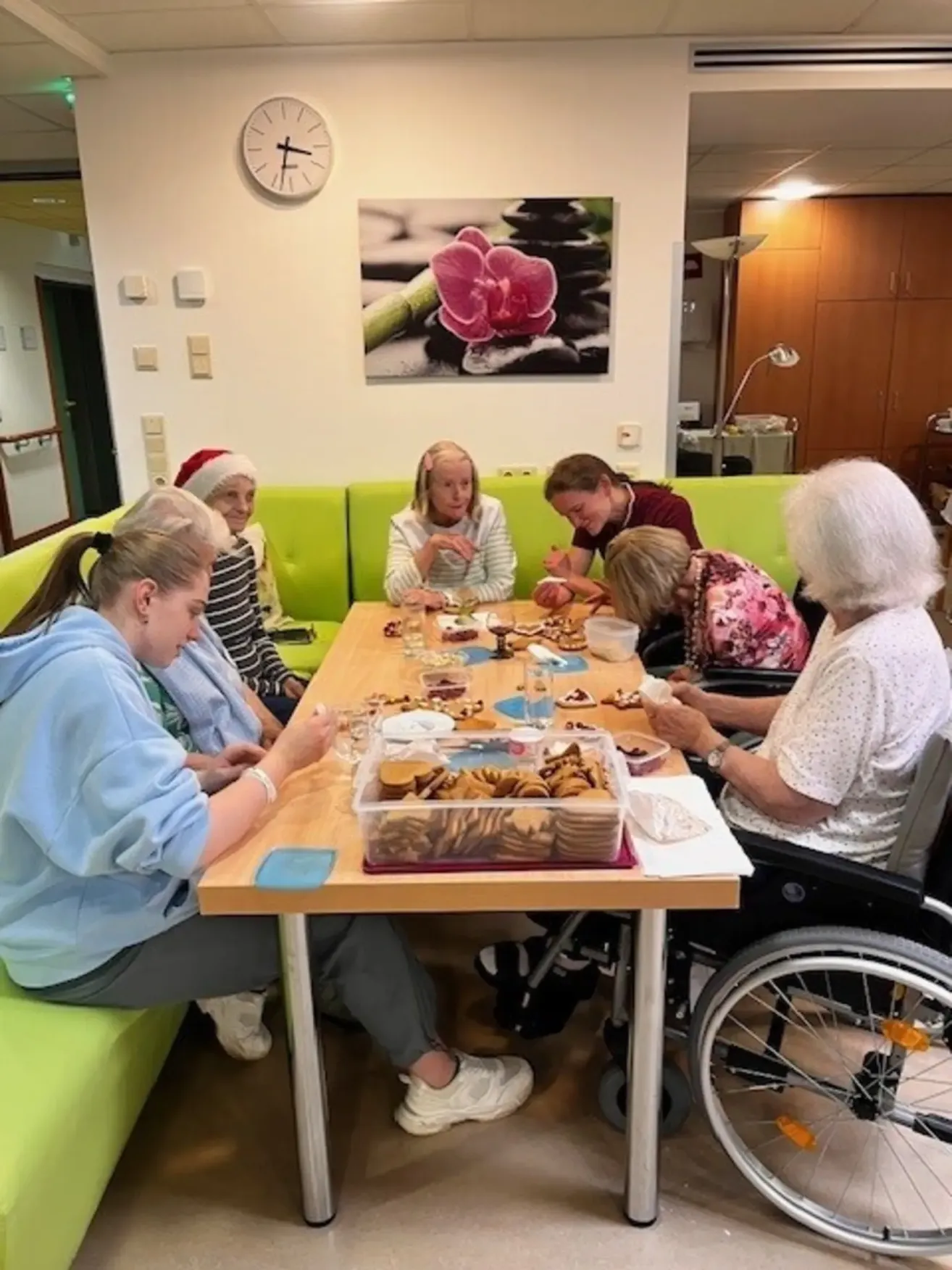 Eine Gruppe älterer Frauen, einige in Rollstühlen, genießen eine Mahlzeit an einem Tisch mit Essen, Wein und Keksen. Dahinter eine Wand mit einer Uhr und einem Gemälde.