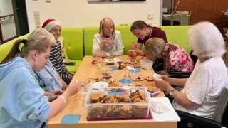 Eine Gruppe älterer Frauen, einige in Rollstühlen, genießen eine Mahlzeit an einem Tisch mit Essen, Wein und Keksen. Dahinter eine Wand mit einer Uhr und einem Gemälde.