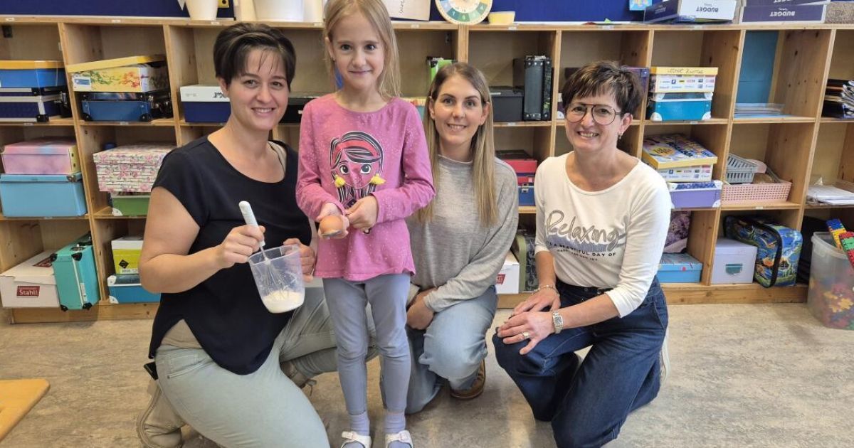 Vier Frauen posieren mit einem kleinen Mädchen, das ein Ei hält, für ein Foto. Sie lächeln und knien auf dem Boden. Dahinter steht ein Holzregal mit Büchern und anderen Gegenständen.