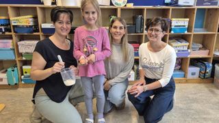 Vier Frauen posieren mit einem kleinen Mädchen, das ein Ei hält, für ein Foto. Sie lächeln und knien auf dem Boden. Dahinter steht ein Holzregal mit Büchern und anderen Gegenständen.