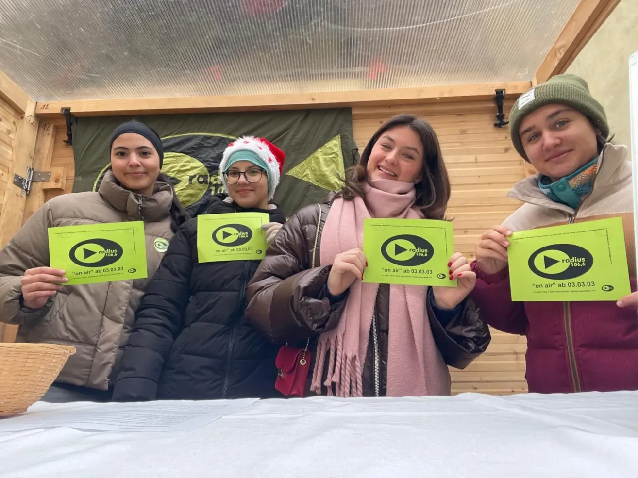 Vier Personen stehen zusammen und halten grüne Schilder mit einem Play-Button und 'on air' darauf. Sie lächeln und scheinen sich bei einer öffentlichen Veranstaltung zu befinden.