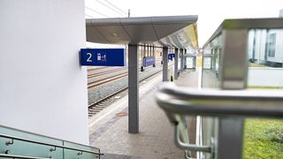 Ein Blick auf einen Bahnhof mit einem blauen Schild, das die Bahnsteignummer '2' anzeigt. Der Bahnsteig ist leer, mit einem Metallgeländer an der Seite.