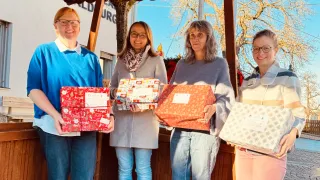 Drei Frauen stehen vor einem Gebäude mit Weihnachtsdekoration und halten verpackte Geschenke. Eine hält eine große rote Box, eine andere eine bunte kleinere Box und die dritte eine kleinere rote Box. Dahinter ein Gebäude mit einem teilweise sichtbaren Schild.