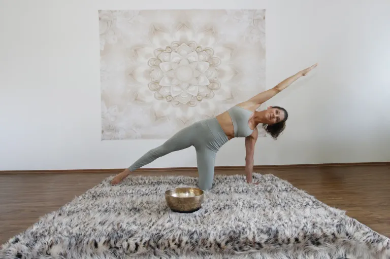 Eine Frau in Trainingskleidung praktiziert Yoga auf einem pelzigen Teppich und nimmt eine Pose mit erhobenen Armen ein. Hinter ihr befindet sich eine Wand mit einem großen weißen Blumendesign.