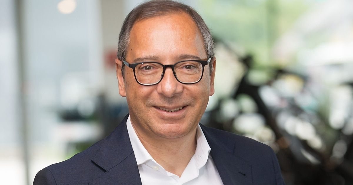 Ein Mann mit grauem Haar und Brille lächelt und schaut in die Kamera. Er trägt ein weißes Hemd und einen dunkelblauen Blazer. Der Hintergrund ist unscharf mit einer Mischung aus grünen und weißen Farben.