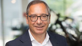 Ein Mann mit grauem Haar und Brille lächelt und schaut in die Kamera. Er trägt ein weißes Hemd und einen dunkelblauen Blazer. Der Hintergrund ist unscharf mit einer Mischung aus grünen und weißen Farben.