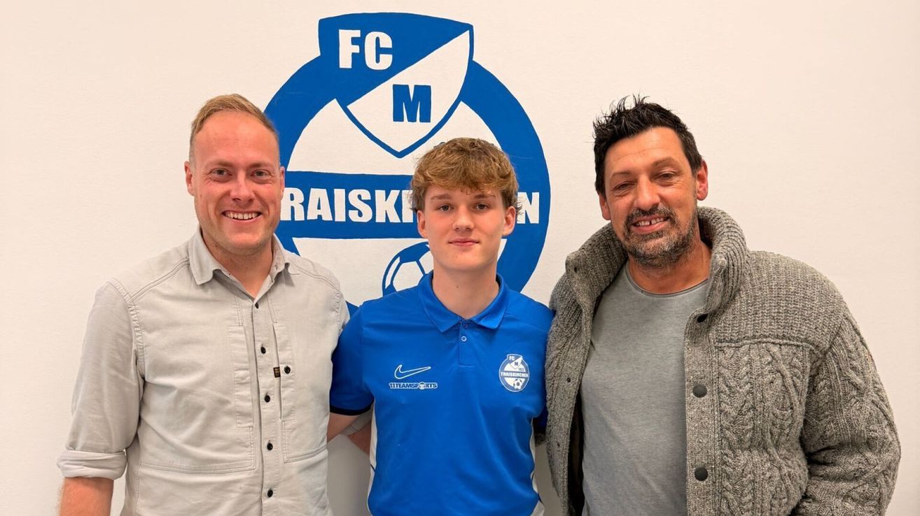 Drei Männer stehen lächelnd zusammen vor einem blauen Logo an einer weißen Wand. Der Mann in der Mitte trägt ein blaues Poloshirt mit dem Nike-Logo.