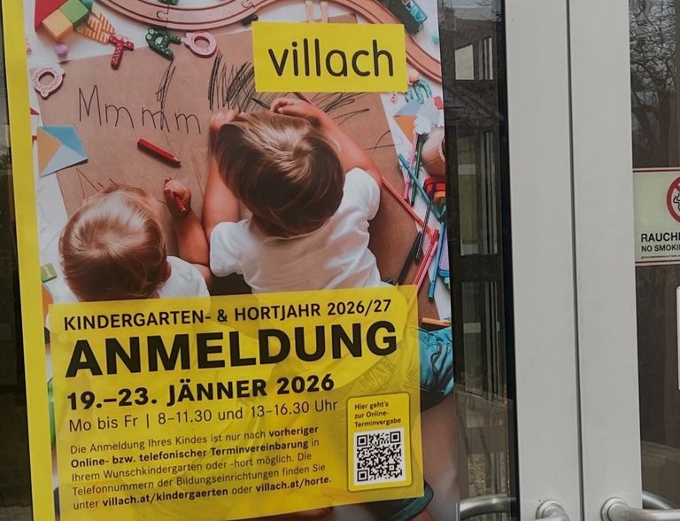 Ein Poster mit zwei kleinen Kindern, die auf einem Pappkarton zeichnen. Der Text auf dem Poster lautet 'VILLACH', 'ANMELDUNG' und '9.-23. JÄNNER 2026'. Es enthält auch einen QR-Code. Die Kinder scheinen beschäftigt zu sein, auf dem Karton zu zeichnen oder zu schreiben.