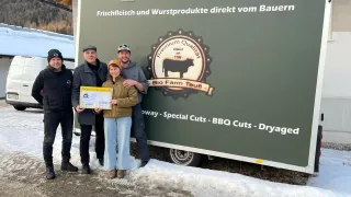 Fünf Personen stehen vor einem Lastwagen. Eine Frau hält eine Urkunde. Der Lastwagen hat einen grünen Hintergrund und weiße Text, der 'Premium Qualität Bio Farm Teufel - Special Cuts - BBQ Cuts - Dryaged' lautet.
